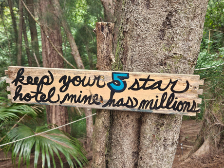 A wooden sign on a tree reads 'Keep your 5 star hotel mine has millions!', an ode to nature's luxury for an RV rental escape.