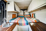 RV rental camper van interior featuring a cozy bed with colorful bedding, a sink, and wooden counters.