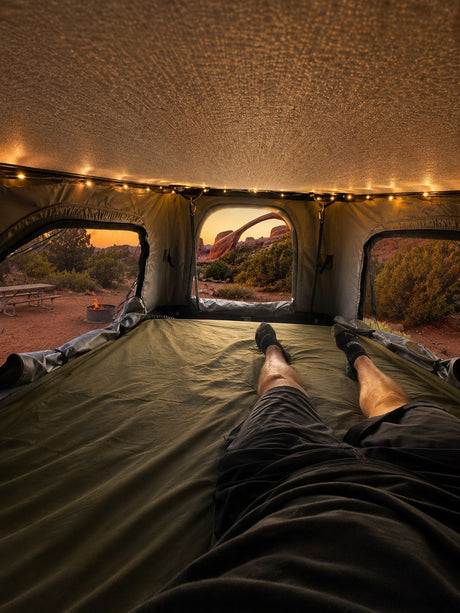 First-person view inside a comfortable rv rental rooftop tent, showing legs of a person and a stunning sunset over a natural arch.