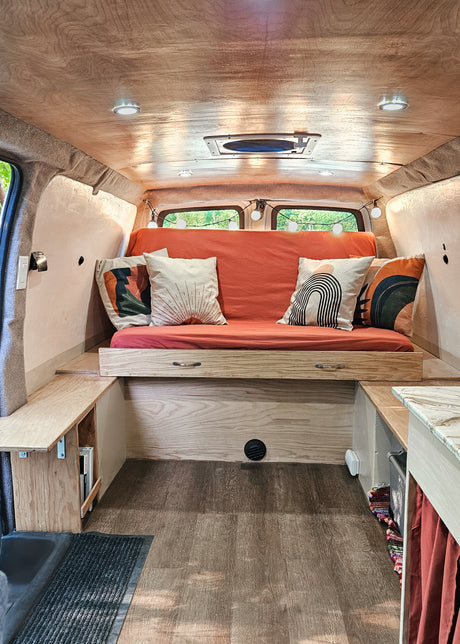 This cozy rv rental camper van interior features a wooden ceiling, comfy seating/bed, storage, & built-in lights.