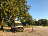 An RV rental camper van, a beige Mercedes Sprinter, parked at a shaded campground site with an extended awning and picnic table.
