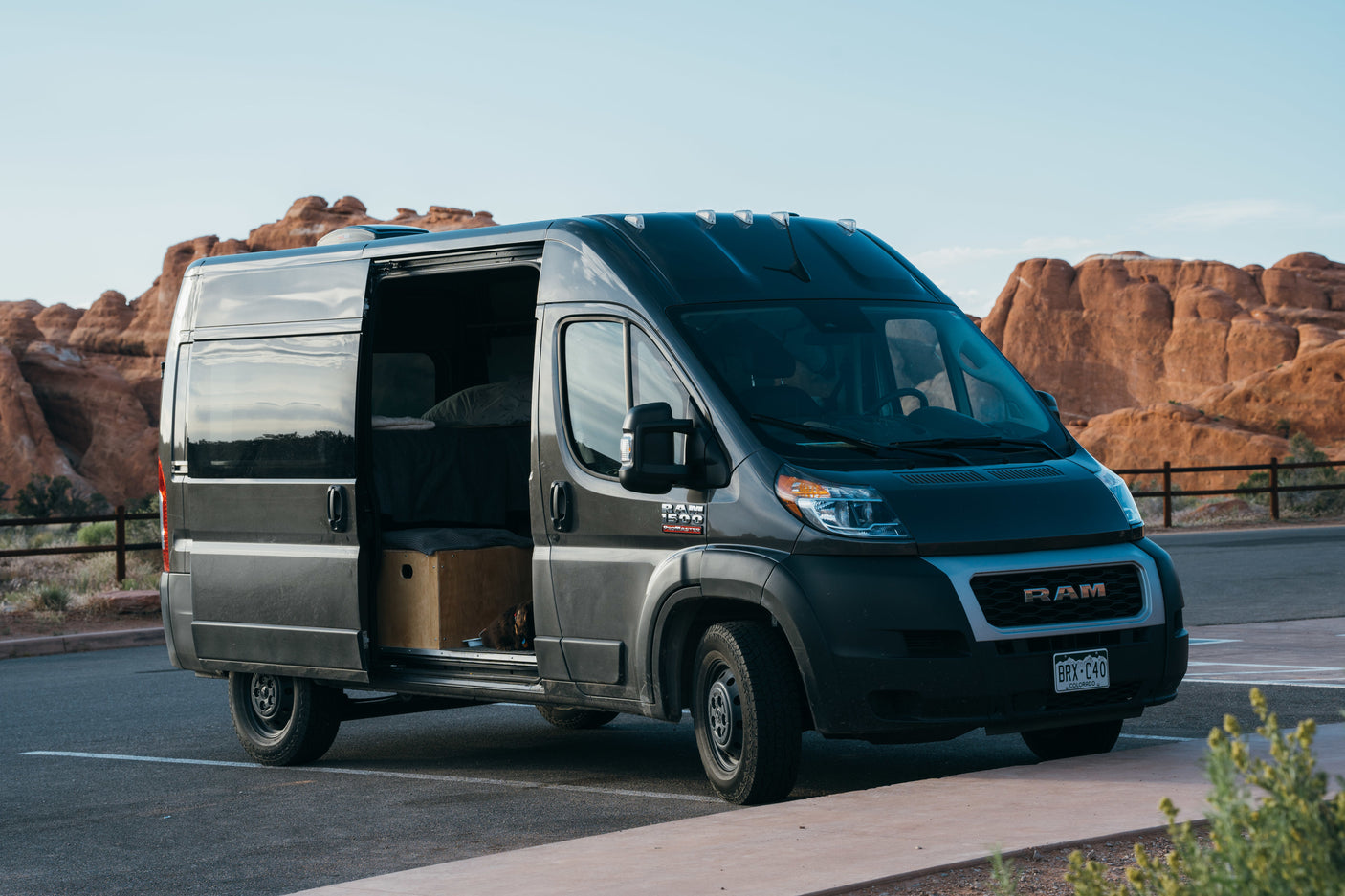 Camper van RV rental: A dark gray Ram ProMaster with its side door open, revealing a bed and dog, parked in a desert landscape.