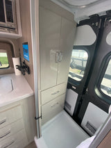 RV rental interior: Compact kitchen with microwave, sink, and tablet. Adjacent storage cabinets and a shower pan are visible.