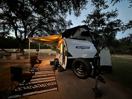 Nighttime rv rental setup: a small teardrop trailer with illuminated awning, camping chairs, and a rooftop tent. Perfect for adventure.