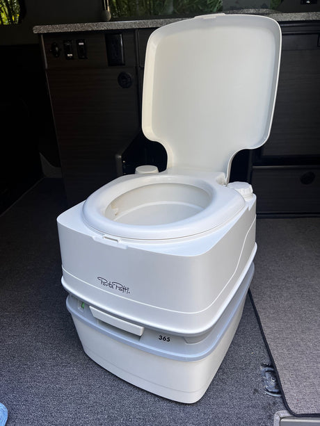 Portable Porta Potti toilet in an RV rental, essential for camping and travel. The white unit sits on dark carpet near wood cabinets.