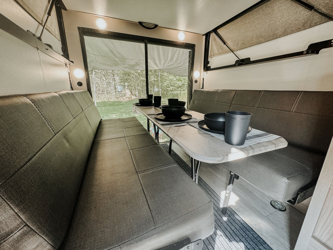 This RV rental's inviting interior features a dinette with benches, a set table, and large windows revealing a forest view.