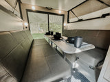 This RV rental's inviting interior features a dinette with benches, a set table, and large windows revealing a forest view.