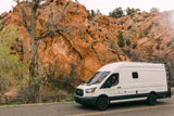 White rv rental van on a paved road with a driver, set against vibrant red rock cliffs and diverse trees.