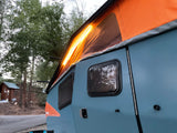 An illuminated Taxa Cricket RV rental with its orange pop-up roof, parked at a rustic campsite among trees.