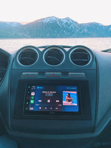 Scenic RV rental view: snowy mountains, a lake, and The National playing on the car's infotainment screen during a winter road trip.