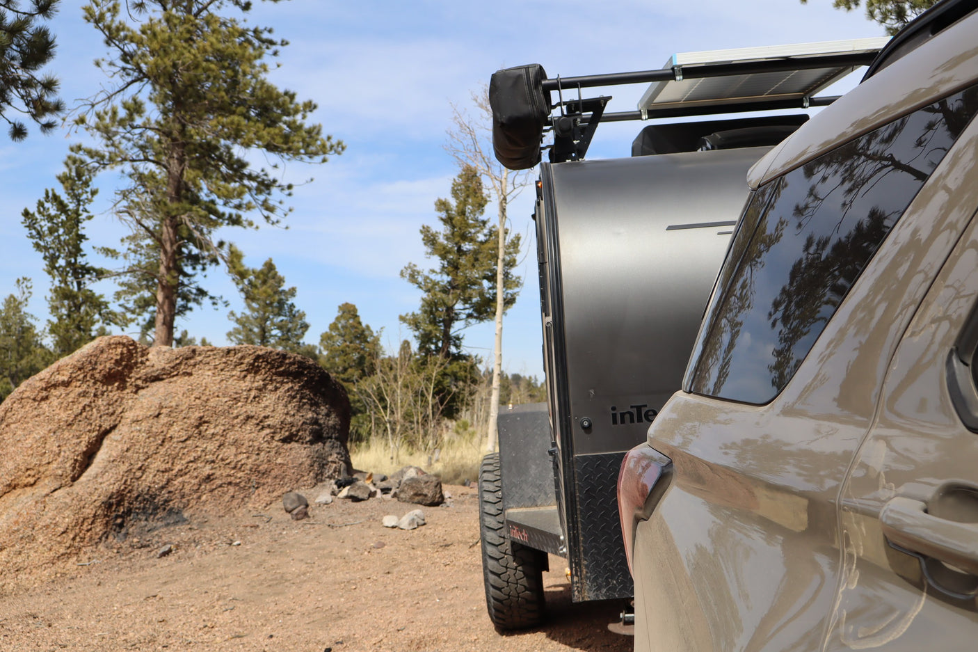 Adventure-ready RV rental: A tan SUV towing a dark grey off-road trailer with solar panels, parked by a rock and pine trees.