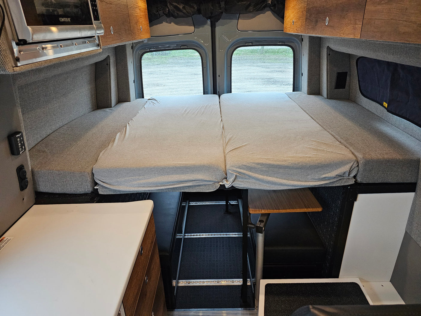 RV rental camper van interior featuring a comfortable convertible bed, microwave, and storage. Rear windows offer a view.