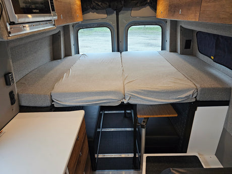RV rental camper van interior featuring a comfortable convertible bed, microwave, and storage. Rear windows offer a view.