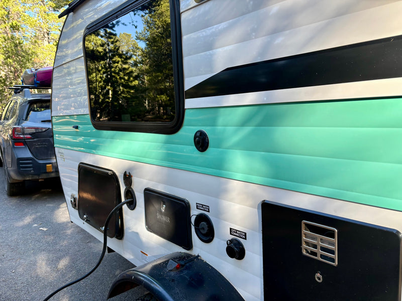 Side of an RV rental with visible utility hookups (power, water, shower), parked next to a car with a kayak, amidst trees. — photo 8