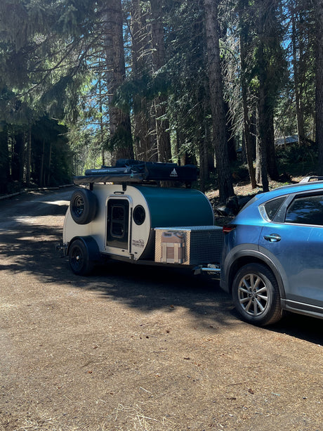An RV rental teardrop camper hitched to a blue SUV on a sunny forest road. Perfect for camping adventures.