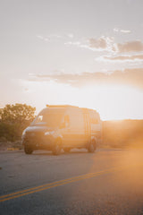 Your next rv rental adventure awaits! A dark Mercedes Sprinter camper van, customized for travel, glows golden on a desert road at sunset.