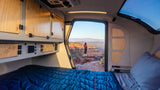 Enjoy the scenic outdoors from this RV rental! A cozy bed inside overlooks a person on a cliff with red rock views.