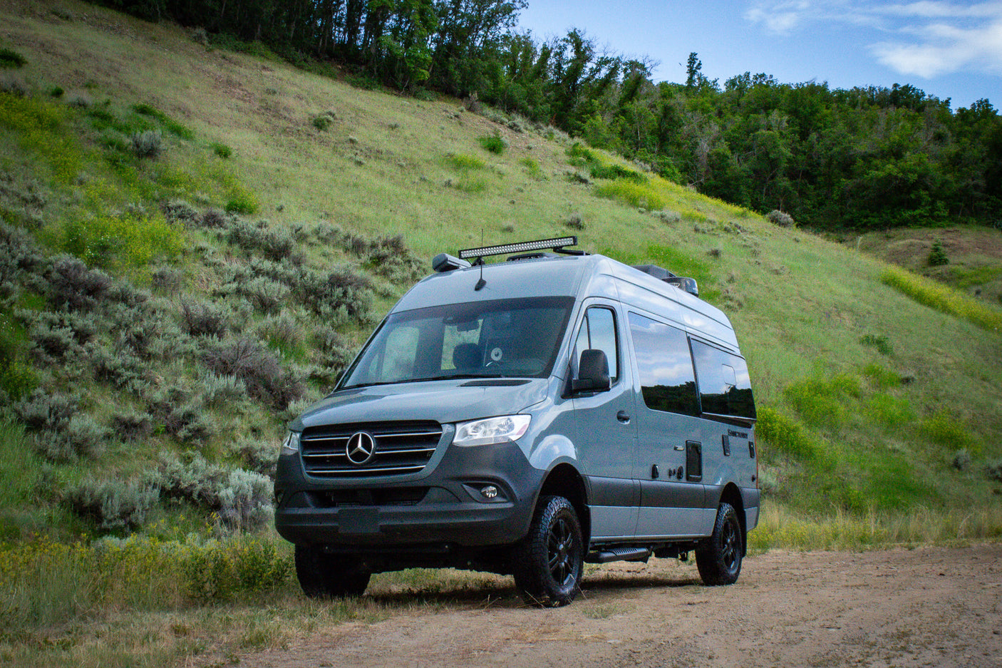Adventure-ready gray RV rental, a Mercedes Sprinter van, parked on a dirt road against green hills.