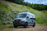 Adventure-ready gray RV rental, a Mercedes Sprinter van, parked on a dirt road against green hills.