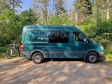 Green Mercedes Sprinter RV rental van with bikes parked on a forest dirt road, ready for adventure.