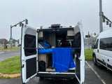 This RV rental shows its open rear doors revealing a cozy bed with galaxy bedding and a blue towel. Parked by Oregon State Parks.
