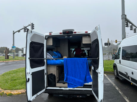 This RV rental shows its open rear doors revealing a cozy bed with galaxy bedding and a blue towel. Parked by Oregon State Parks.