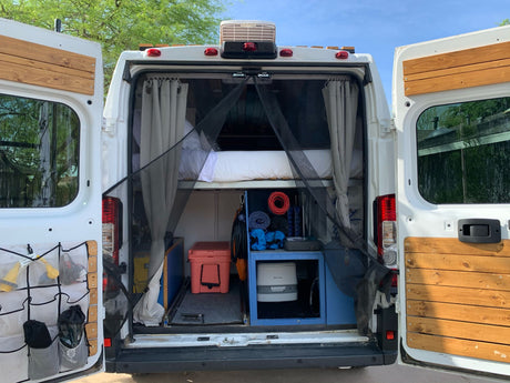 Rear view of an RV rental: a white camper van with open doors showcasing a bed, storage, and compact living space. Ready for trips!