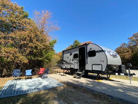 A grey RV rental, a Wolf Pup travel trailer, sits leveled at a scenic autumn campsite with outdoor chairs and a rug.