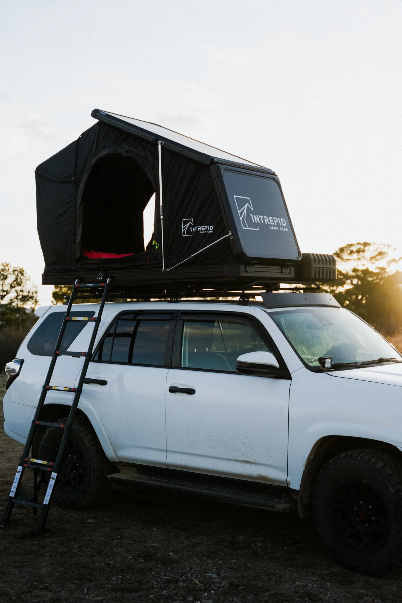 Intrepid Camp Gear Geo Solo hardshell rooftop tent open in wedge position on a roof rack atop a white Toyota 4Runner at sunset, showing the black canopy with front and side entry, the aluminum shell with the Intrepid Camp Gear logo, and the telescoping ladder down to the ground — photo 1