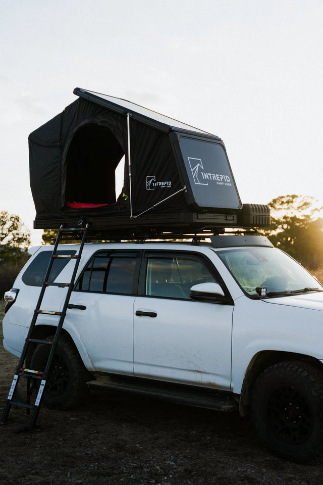 Intrepid Camp Gear Geo Solo hardshell rooftop tent open in wedge position on a roof rack atop a white Toyota 4Runner at sunset, showing the black canopy with front and side entry, the aluminum shell with the Intrepid Camp Gear logo, and the telescoping ladder down to the ground