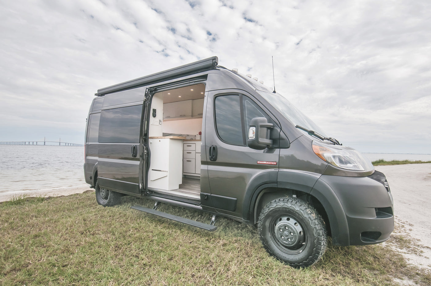 Custom grey Ram ProMaster camper van (RV rental) by water, door open revealing kitchen. Ready for travel adventure.