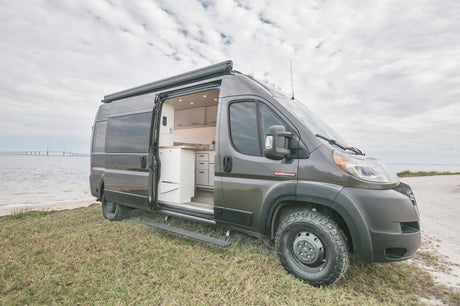 Custom grey Ram ProMaster camper van (RV rental) by water, door open revealing kitchen. Ready for travel adventure.