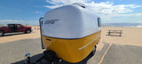 A white & yellow Scamp travel trailer (RV rental) and red truck are parked by a sandy beach with a picnic table & ocean.
