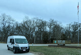 White rv rental camper van at Beaver Dam State Park entrance, with bare trees, park sign, and US & state flags under a cloudy sky.