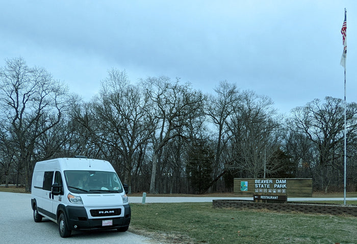 White rv rental camper van at Beaver Dam State Park entrance, with bare trees, park sign, and US & state flags under a cloudy sky.