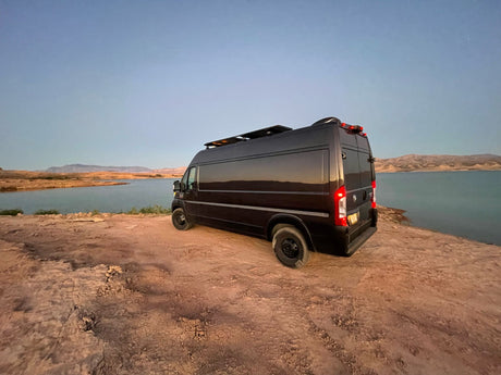 Black camper van, an ideal RV rental, parked lakeside on dirt. Distant mountains under a blue sky, perfect for outdoor adventure.