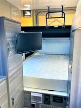 Interior of an RV rental showing a compact bedroom with a bed, wall-mounted TV, cabinets, & control panels.