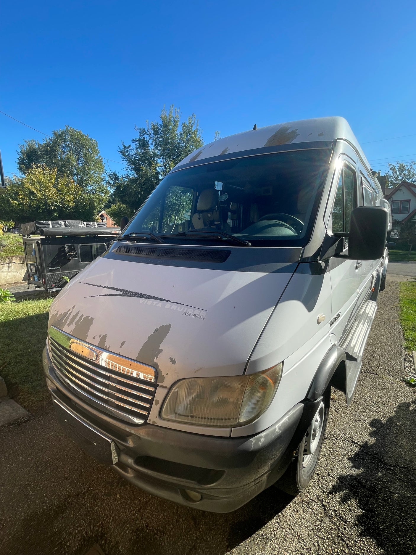 Sunny day view of a grey Vista Cruiser RV rental van, showing paint wear, parked on a driveway with an overland trailer nearby.