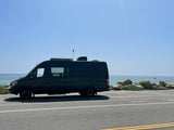 Dark green RV rental van parked by the sparkling blue ocean on a sunny day, with a distant sailboat. Scenic travel.