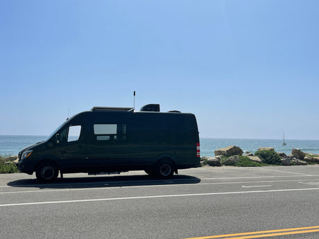 Dark green RV rental van parked by the sparkling blue ocean on a sunny day, with a distant sailboat. Scenic travel.