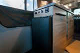 RV rental: Compact kitchen area in a camper van, showing a small fridge, control panel with outlets, and wood cabinets.