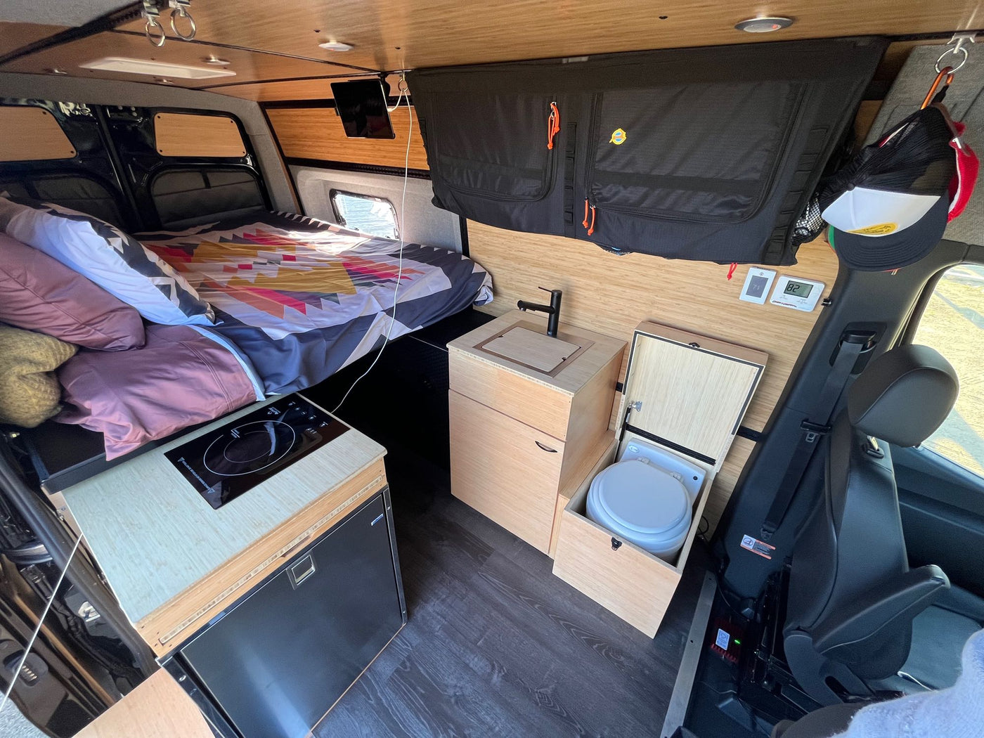 Warm wood tones accent this RV rental's camper van interior: bed, compact kitchen, fridge, and pull-out toilet. — photo 14