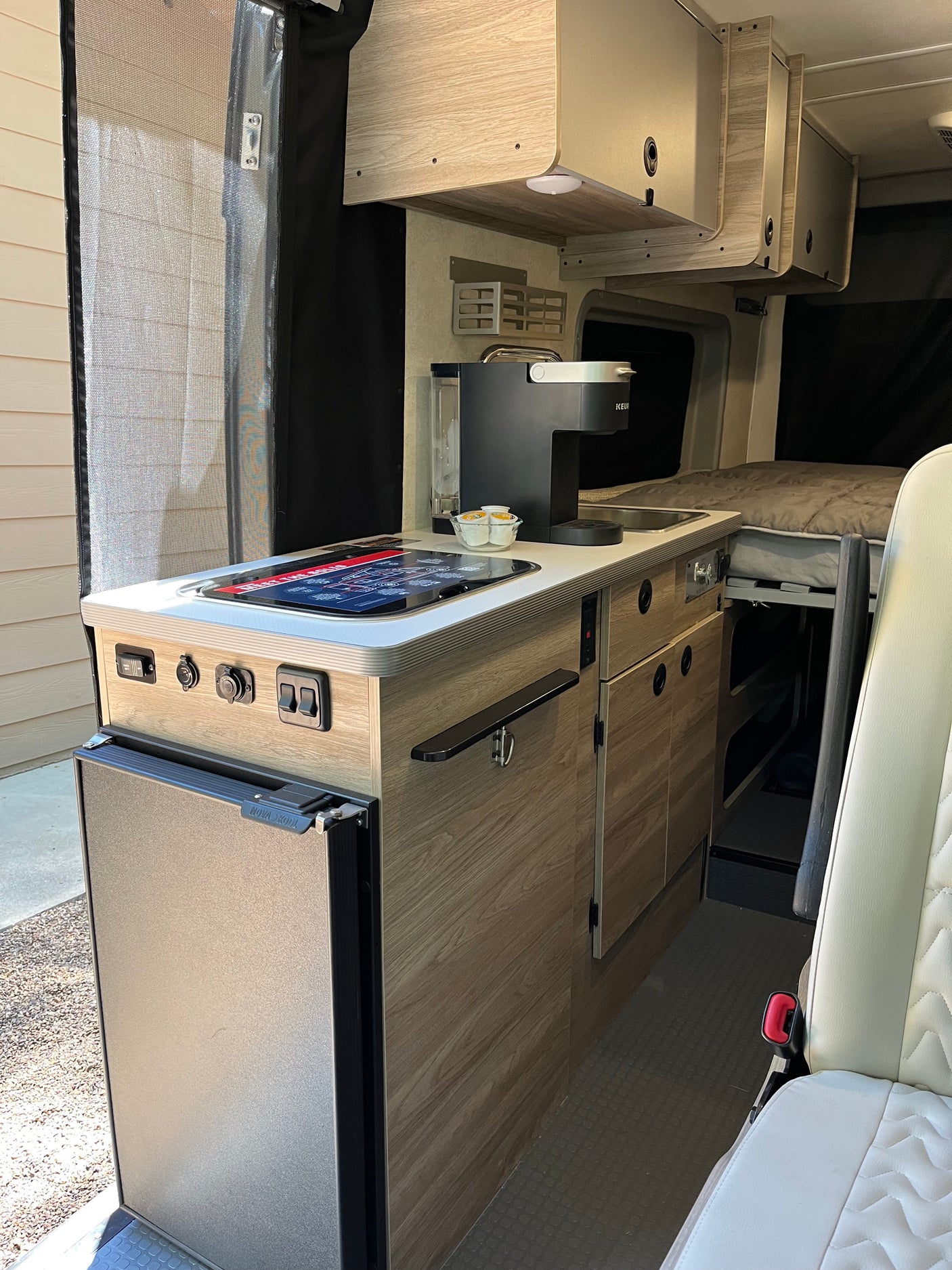 Inside this RV rental, a compact camper van kitchenette with a coffee maker and fridge; a cozy bed is visible beyond.
