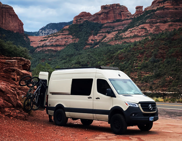 White RV rental camper van with bikes on a red dirt road, set against magnificent red rock mountains.