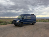 Dark blue Sprinter van RV rental parked on a gravel road, with a cloudy sky overhead and vast, open landscape behind it.