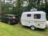 Black SUV towing a white Scamp rv rental travel trailer on a grassy area, surrounded by green trees.