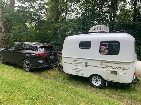 Black SUV towing a white Scamp rv rental travel trailer on a grassy area, surrounded by green trees.