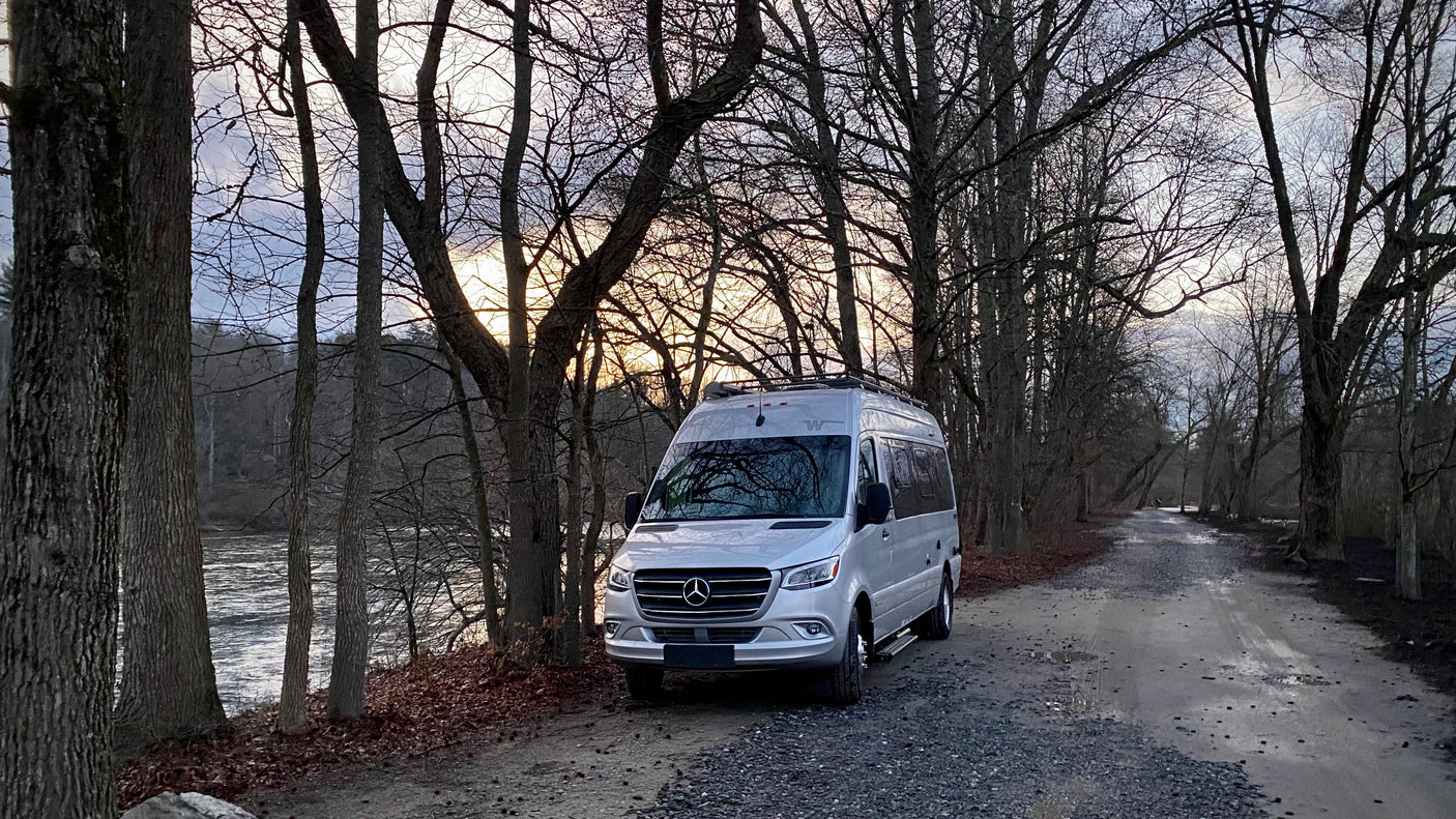 Silver Mercedes RV rental parked by a peaceful river on a gravel road, surrounded by bare trees at sunset.
