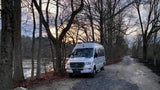 Silver Mercedes RV rental parked by a peaceful river on a gravel road, surrounded by bare trees at sunset.