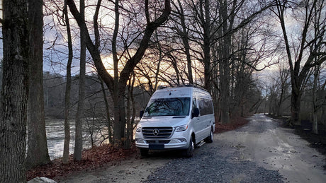 Silver Mercedes RV rental parked by a peaceful river on a gravel road, surrounded by bare trees at sunset.